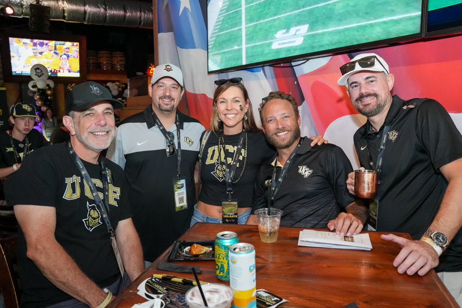 Five friends in UCF apparel pose at a sports bar tailgate table, smiling with drinks and snacks on the counter, large screens in the background.