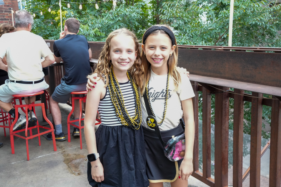Two smiling young girls pose together on a balcony, wearing black and white outfits and festive bead necklaces at an outdoor party.