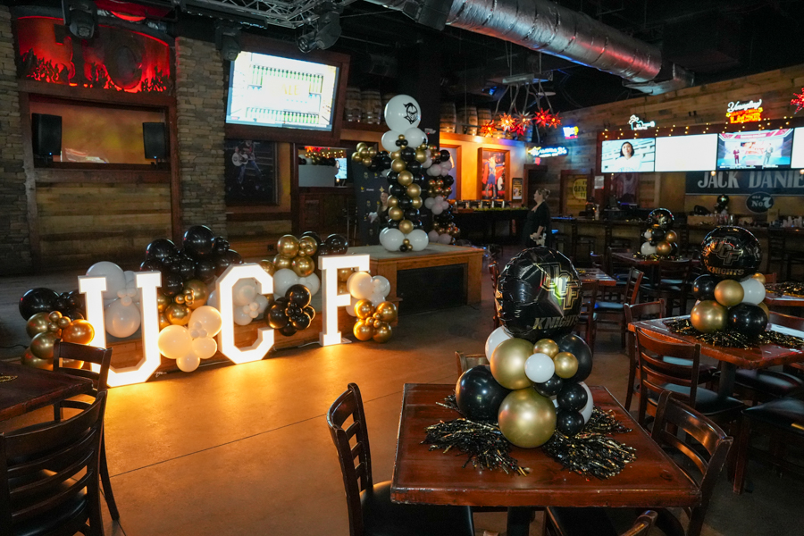 Event space with large illuminated letters 'JCF' and black, white, and gold balloon clusters along a wooden stage/place setting for a celebration.