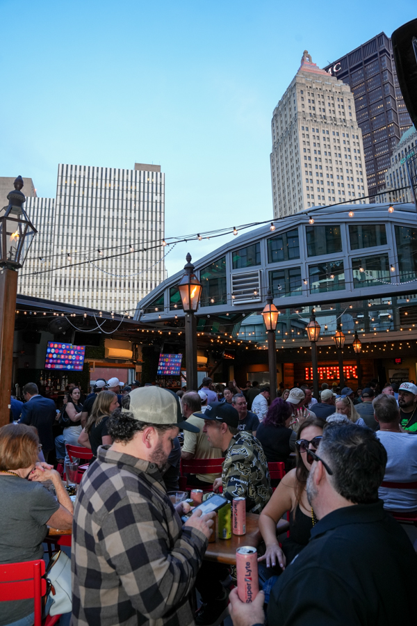 Crowded rooftop bar in a city with string lights, red chairs, and a glass atrium, tall skyscrapers in the background at dusk/early evening.