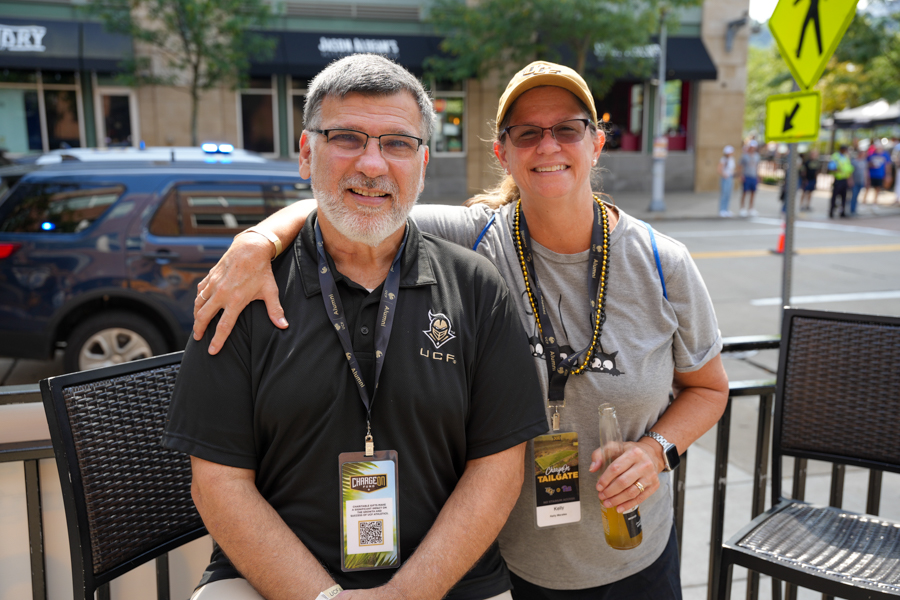 Two smiling adults sit arm-in-arm at an outdoor cafe on a city street, wearing event lanyards.