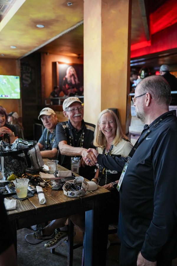 Man in a black jacket shakes hands with a guest across a bar, smiles all around, casual gathering in a dimly lit venue.