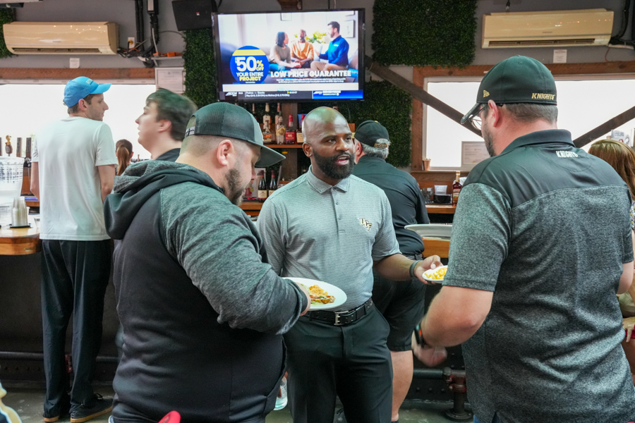 Group of men at a bar; one in a gray UCF polo holds a plate while chatting with two others who also have plates of food.