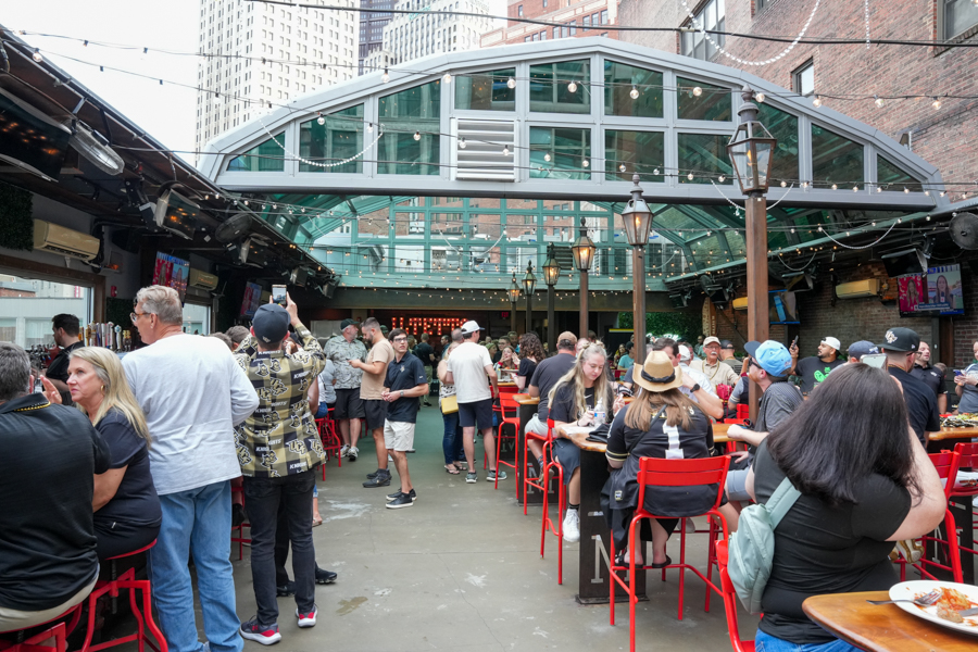 Busy outdoor beer garden with red stools and string lights under a glass-roofed structure, people socializing and eating.