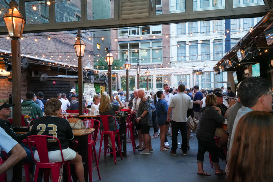 Crowded outdoor patio with string lights and red chairs, people chatting and mingling at dusk.