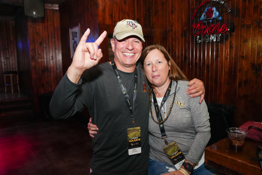 Two people pose together at a tailgate bar; man in a beige cap makes the 'horns' gesture while the woman in a UF gray shirt rests her arm around him.