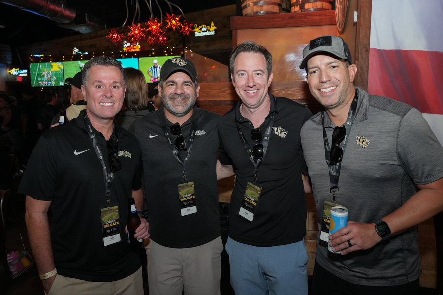 Four men in UF gear pose together at a tailgate party in a bar, smiling for the photo with drinks and lanyards nearby.