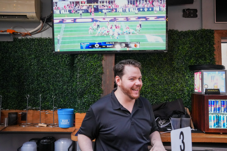 Smiling bartender in a black polo stands in a sports bar with a large TV showing a college football game behind him.