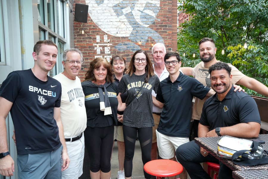 Nine people pose together outdoors on a cafe patio, smiling for the group photo with a brick wall and string lights behind them.
