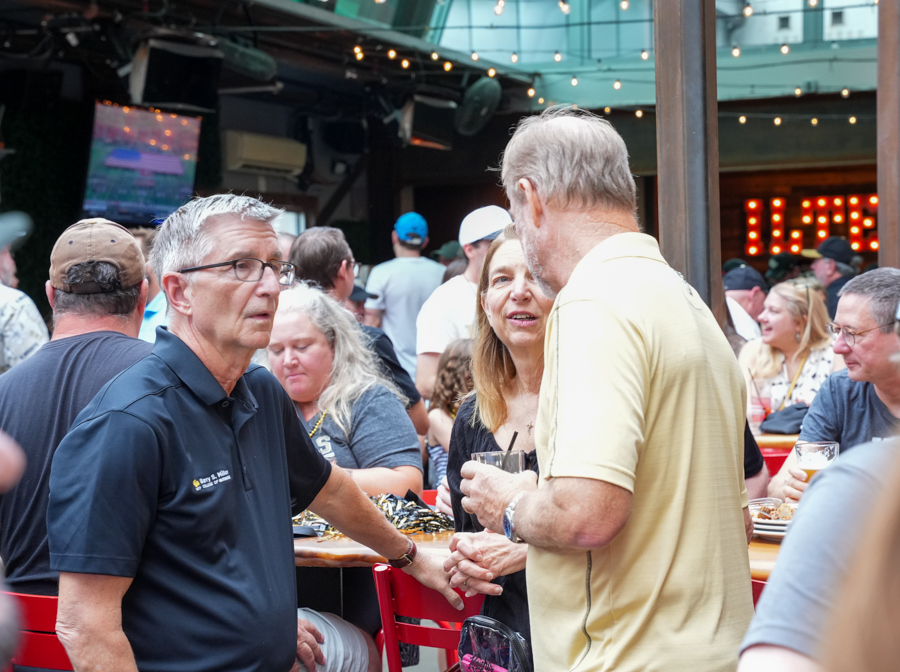 People mingle at a crowded outdoor bar under string lights; two men shake hands while a woman talks nearby, drinks on the table.