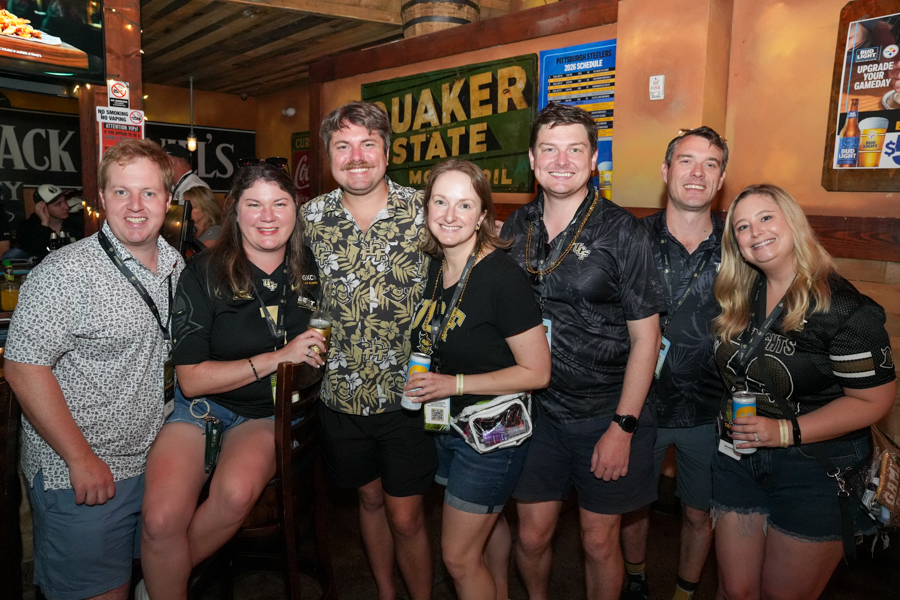 Seven friends posing together in a rustic bar, some holding drinks, with a Quaker State sign and bar decor in the background.