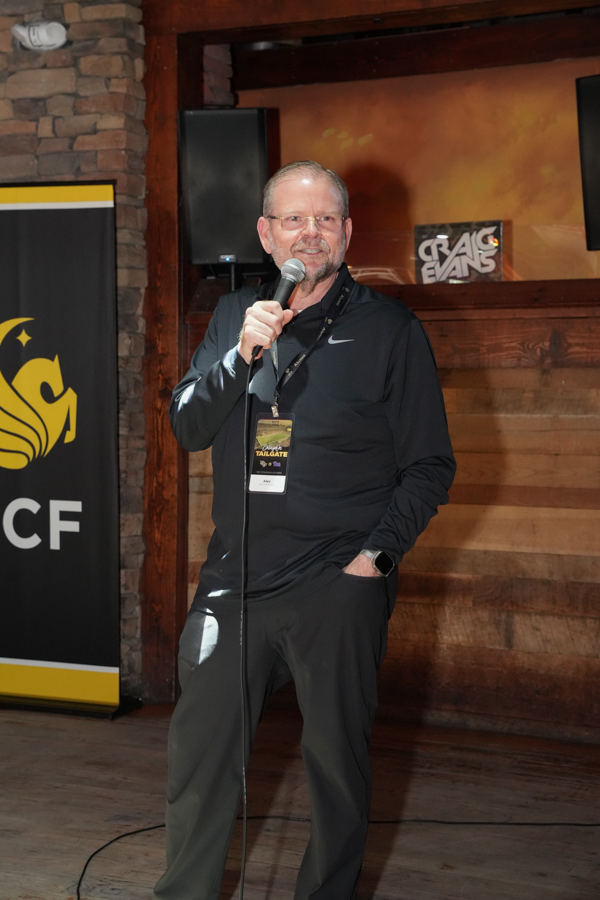 Man on a stage holds a microphone, wearing dark athletic clothing and a lanyard with an ID badge.