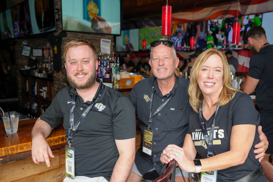 Three people sit at a bar smiling at the camera, wearing dark UCF shirts and lanyards with event badges.