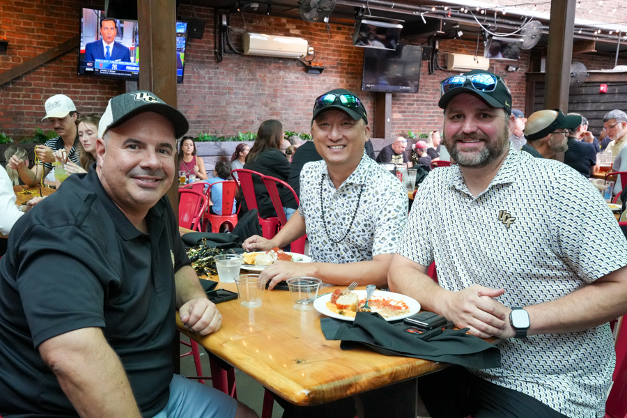 Three men sit at a wooden table in a brick-walled restaurant, smiling at the camera with plates and drinks in front of them.