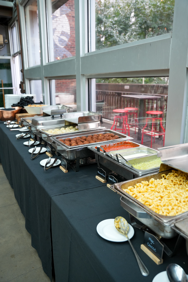 Buffet with multiple hot dishes in chafing pans on a black-clothed table, including pasta and meatballs, beside a windowed wall.