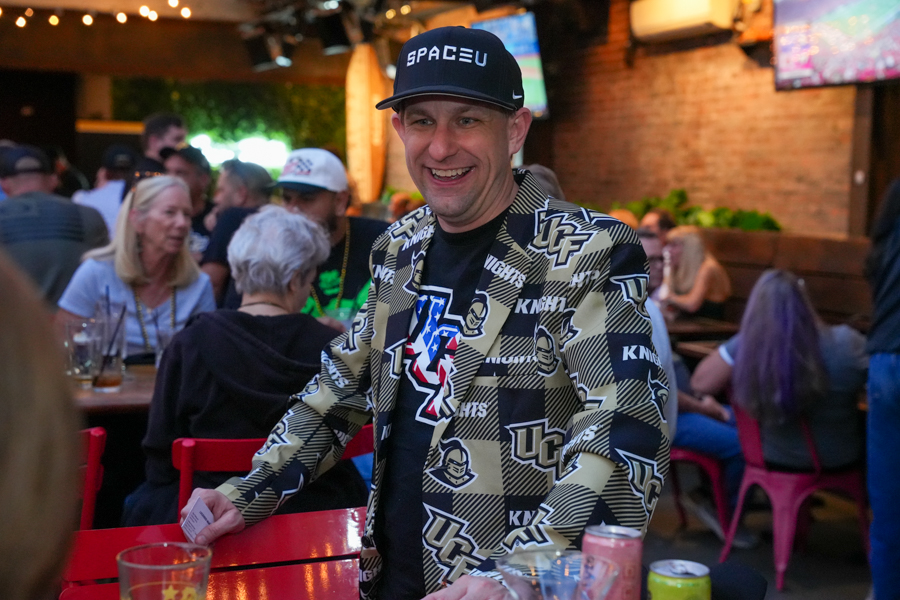 Smiling man wearing a patterned blazer and a black cap at a crowded bar setting near a red table.