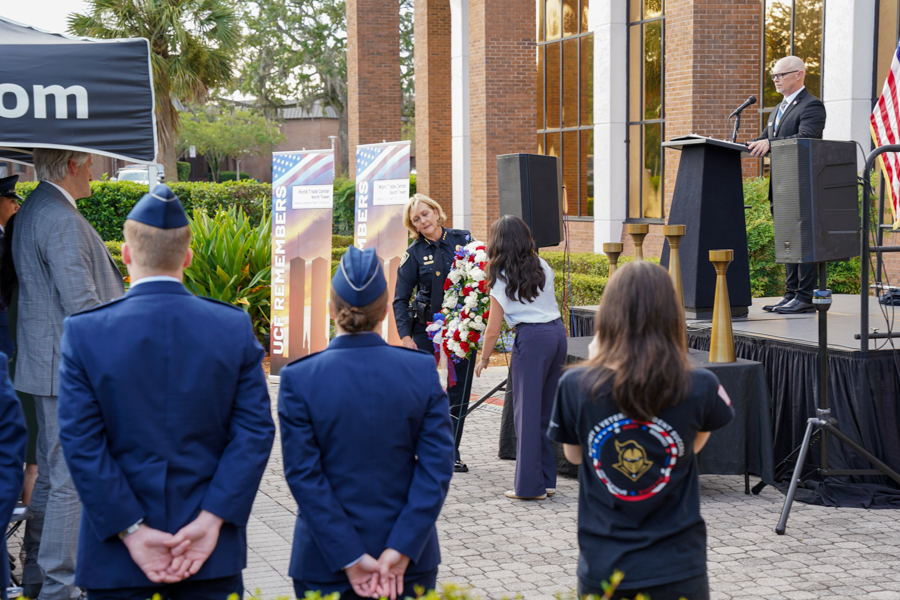 Outdoor memorial ceremony: speaker at podium, a woman in uniform laying a wreath as cadets face the stage; banners and flag in the background.