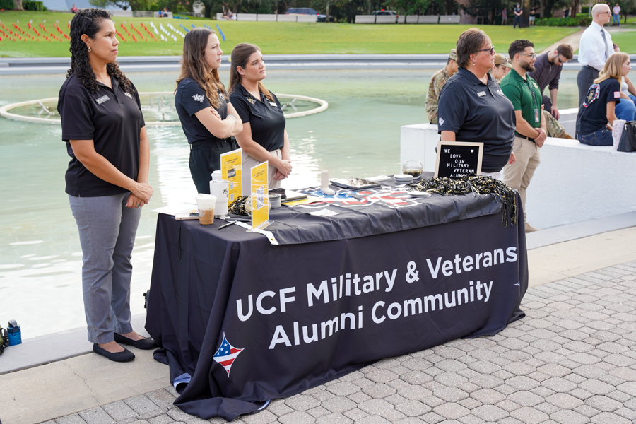 Table promoting UCF Military & Veterans Alumni Community at an outdoor event by a fountain; volunteers stand behind the table with brochures and swag badges visible.