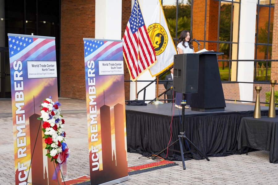 Woman speaks at a memorial event on a stage with a podium, American flag, a university flag, and tall memorial banners reading 'UCF REMEMBERS' and 'World Trade Center North Tower'.