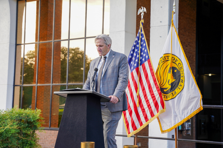 Man in a gray suit speaks at a podium with a microphone outdoors, flanked by the U.S. flag and a university flag behind him.