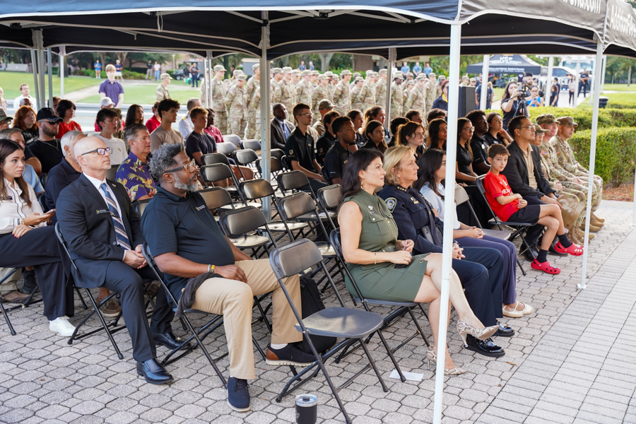 Audience seated under tents at an outdoor ceremony; civilians and service members watch a lineup in the background?