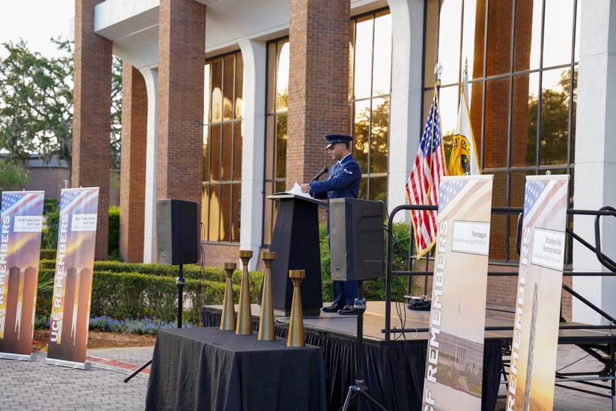 Uniformed speaker delivers a speech at an outdoor ceremony on a stage beside a brick building, with American flag and banners nearby.