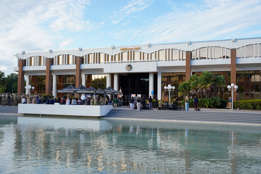 Outdoor ceremony in front of Millican Hall, with a crowd gathered on the steps and a reflecting pool in the foreground.