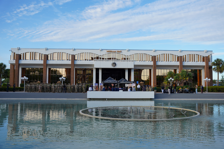 Exterior view of a large civic building with arched windows, a stage, and a crowd; soldiers stand in formation along the steps as civilians gather for a ceremony by a reflecting pool.