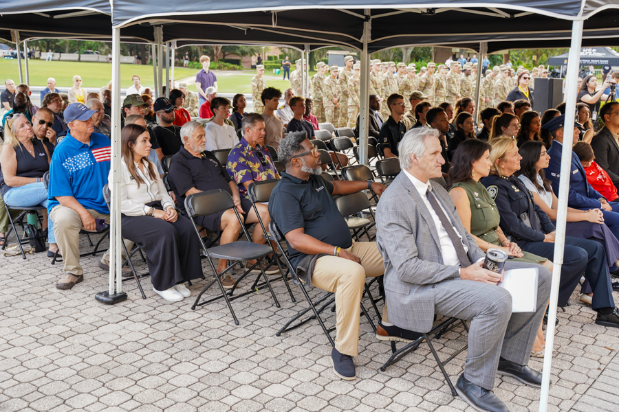 Audience seated under a canopy at an outdoor ceremony, with soldiers in uniform visible in the background.