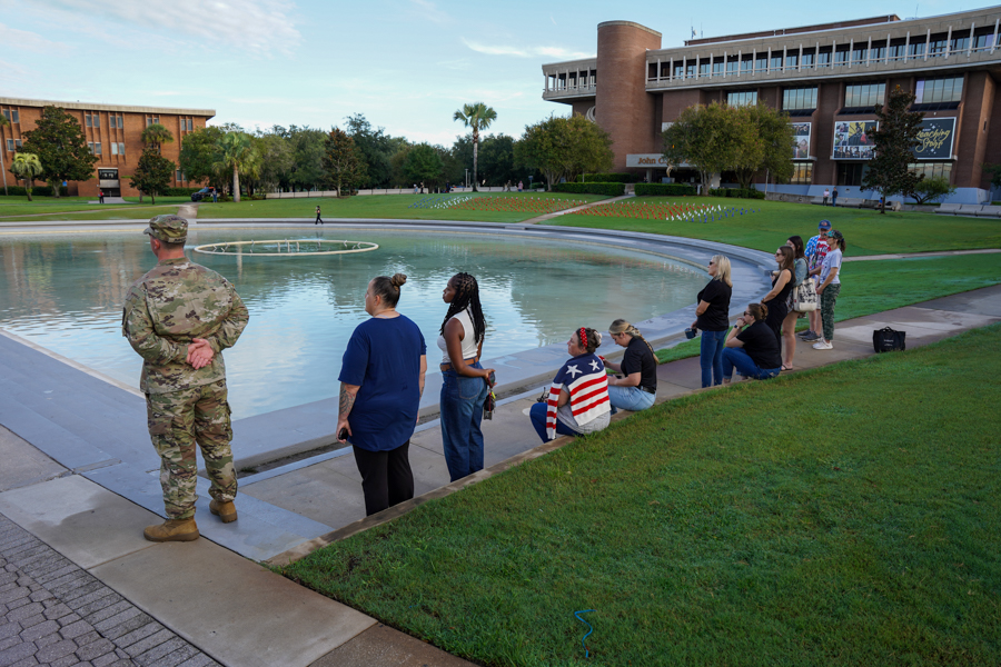 Soldier in camouflage stands with back to camera at edge of a circular fountain, while a group of students sits and stands nearby on a campus quad.