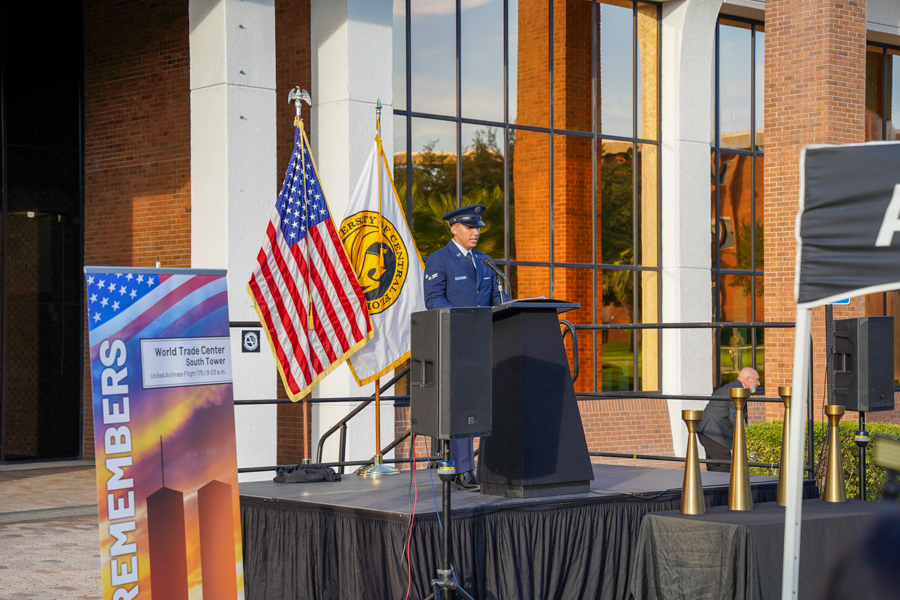 Uniformed man speaks at a podium on an outdoor stage with American and flag banners behind him, near a brick building.