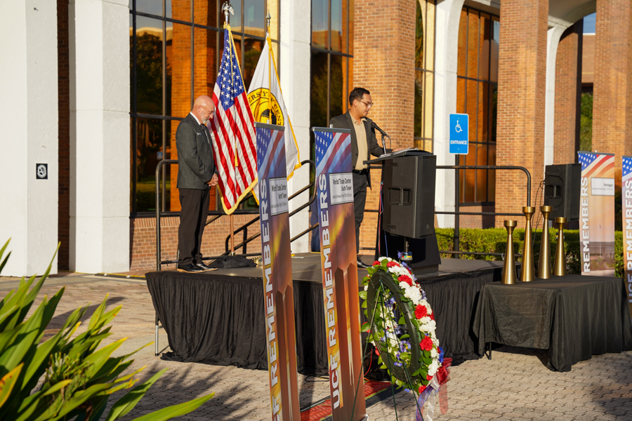 Speaker at a podium on an outdoor stage beside flags and memorial banners, with a wreath nearby in front of a brick building.