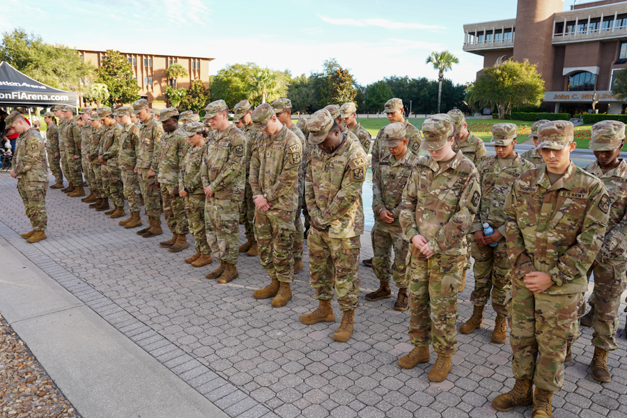 Line of U.S. Army soldiers in camouflage uniforms standing in formation on a paved plaza outdoors. The group faces forward during a ceremony or review.