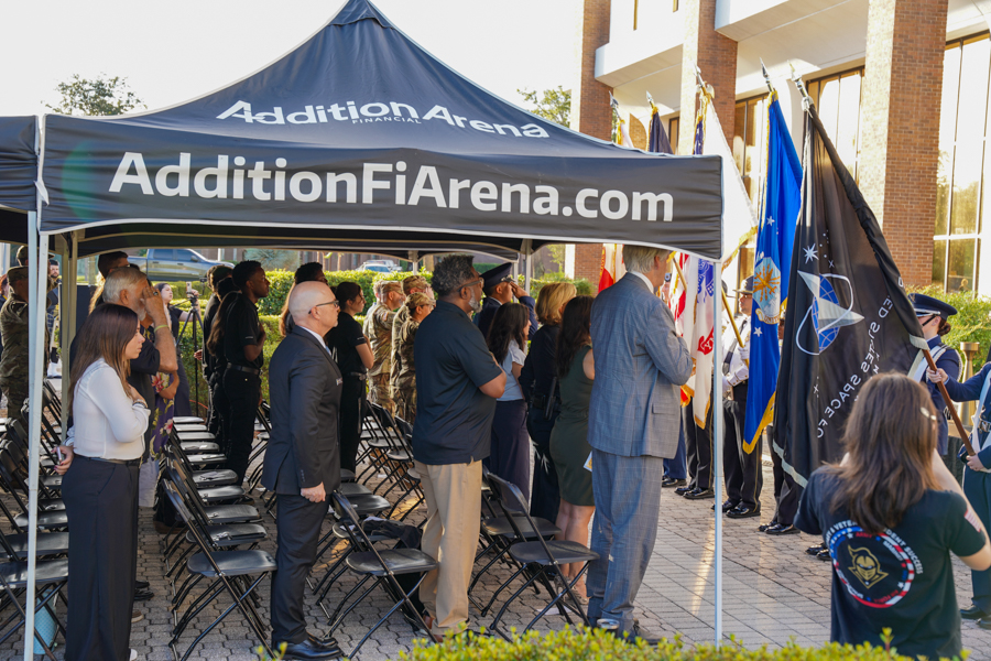 Outdoor ceremony under a blue tent labeled AdditionArena.com with people standing and flags nearby.