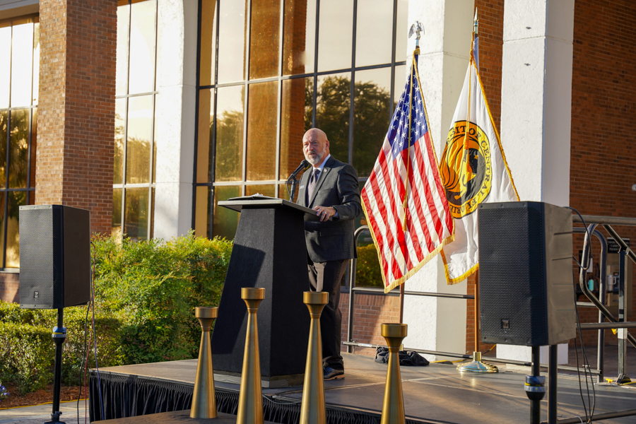 Bearded man in a suit speaking at a lectern on an outdoor stage, flanked by the American flag and a ceremonial flag afterward.