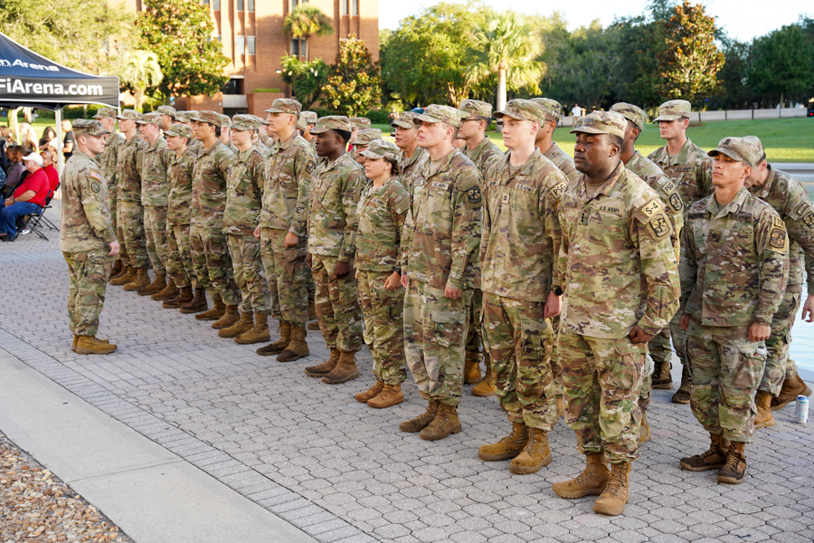 Group of soldiers in desert camouflage standing in formation on a paved outdoor area during a military ceremony, with onlookers and a black tent in the background.