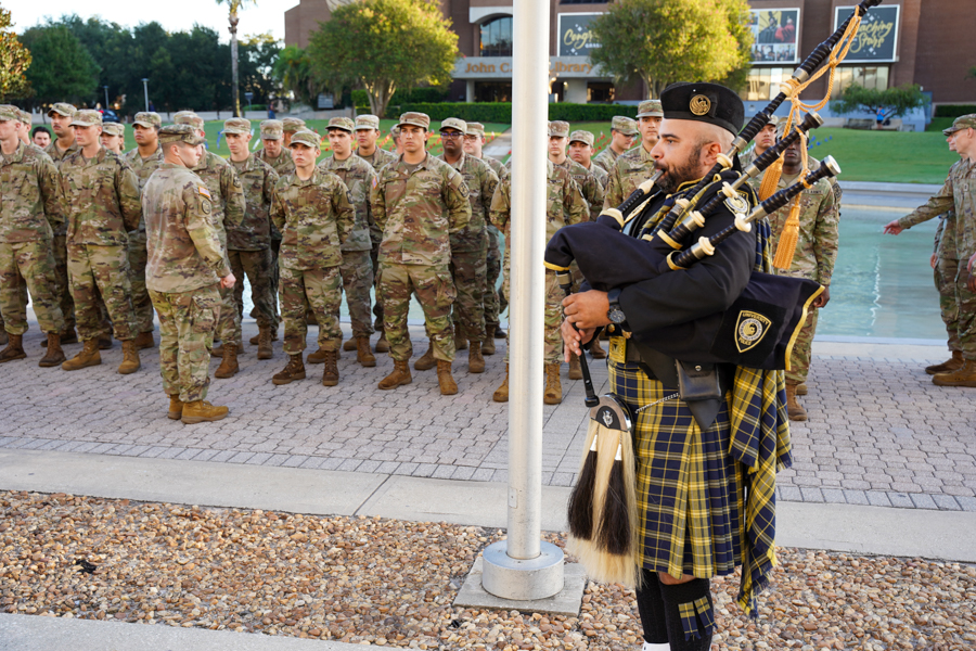 Soldiers in camouflage stand in formation as a bagpiper in a tartan kilt plays in front of them during a ceremony.
