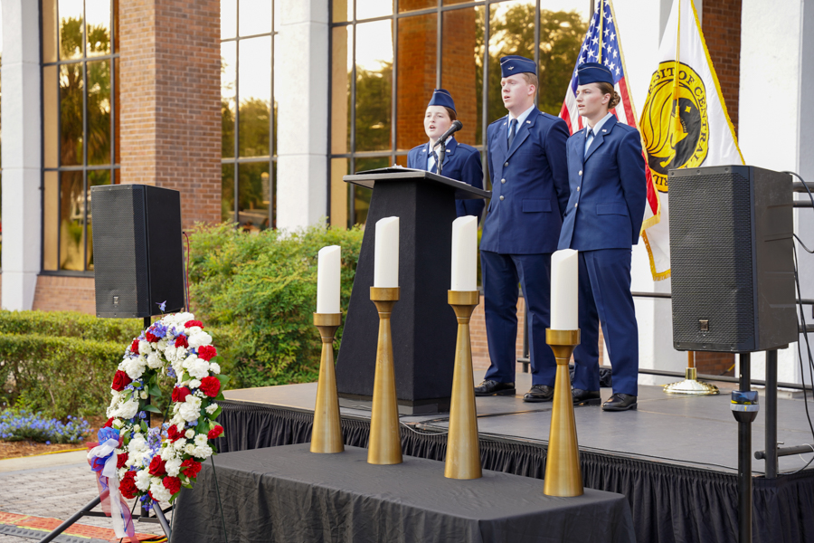 Three uniformed cadets stand at a podium on an outdoor stage, with flags and a wreath nearby.