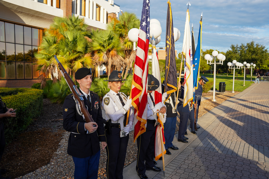 Color guard of uniformed service members standing at attention with multiple flag poles, including the American flag, outside a building.