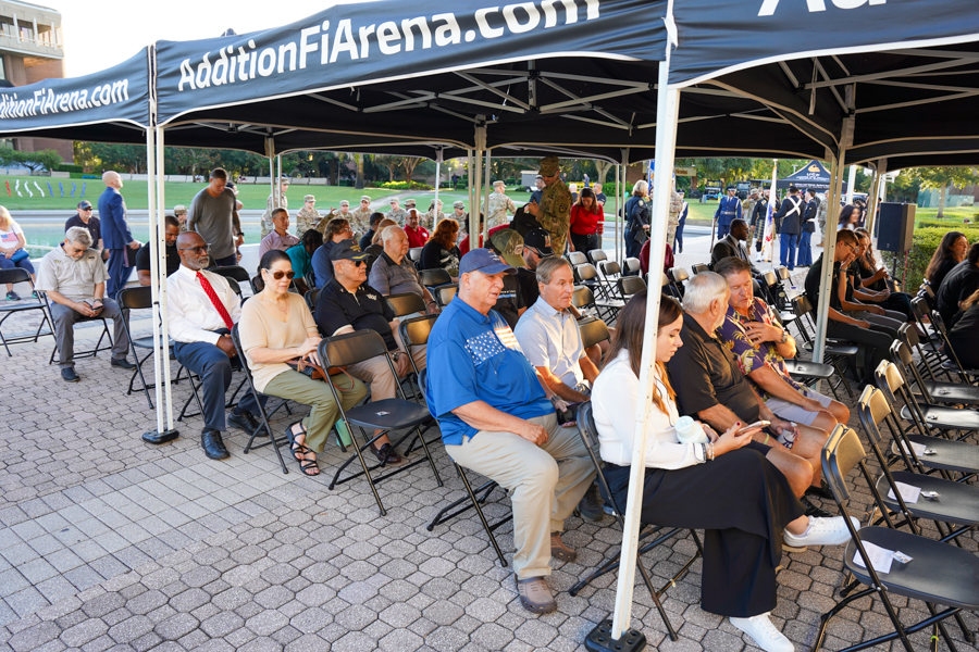 People seated under a blue event canopy at an outdoor ceremony or gathering, with a mix of civilians and veterans in chairs on a paved area.