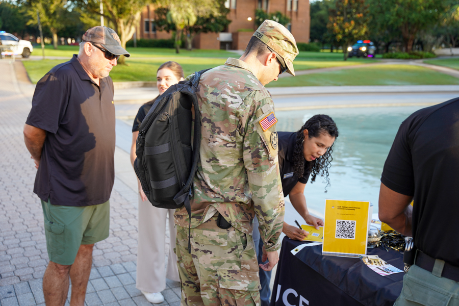 Uniformed service member with backpack talking to civilians at an outdoor community table, a yellow QR-code sign on the table nearby.