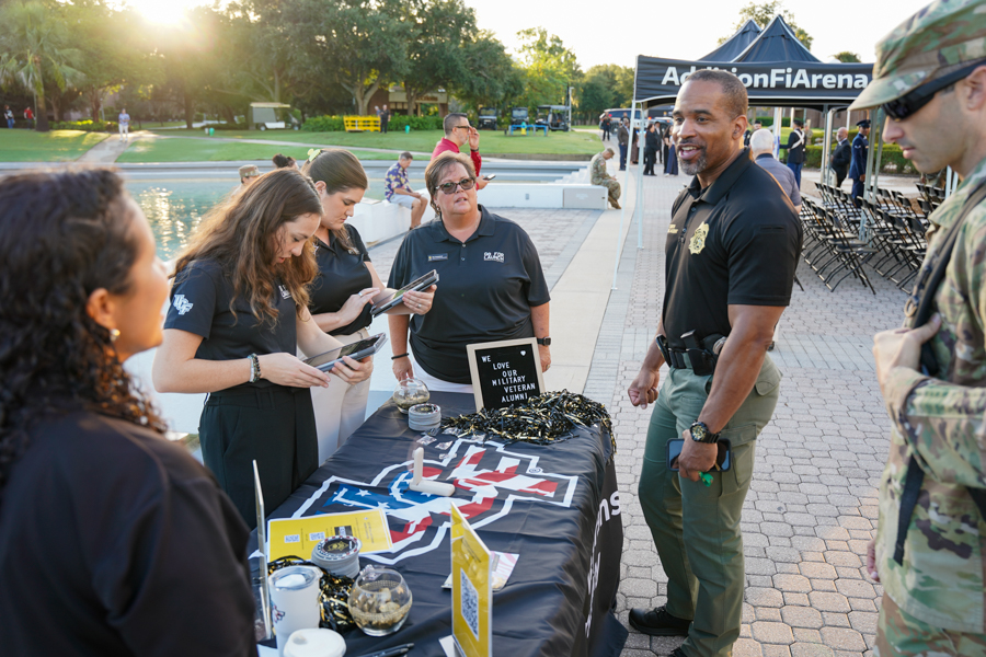 Veterans outreach booth at a poolside event where attendees chat with a uniformed officer and volunteers on laptop displays.