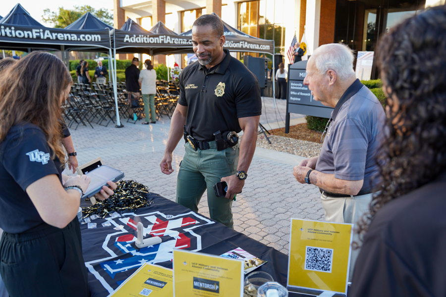 Police officer in black polo interacts with attendees at a table with a UCF-themed banner and yellow info cards outdoors for a college recruitment event