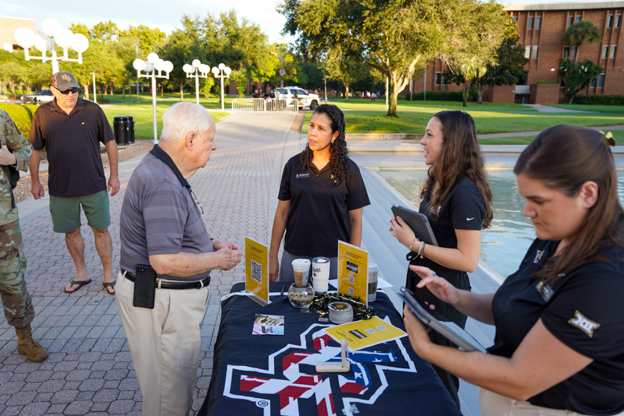 Outdoor campus event: an older man talks with volunteers at a table with yellow flyers and coffee cups on the table.