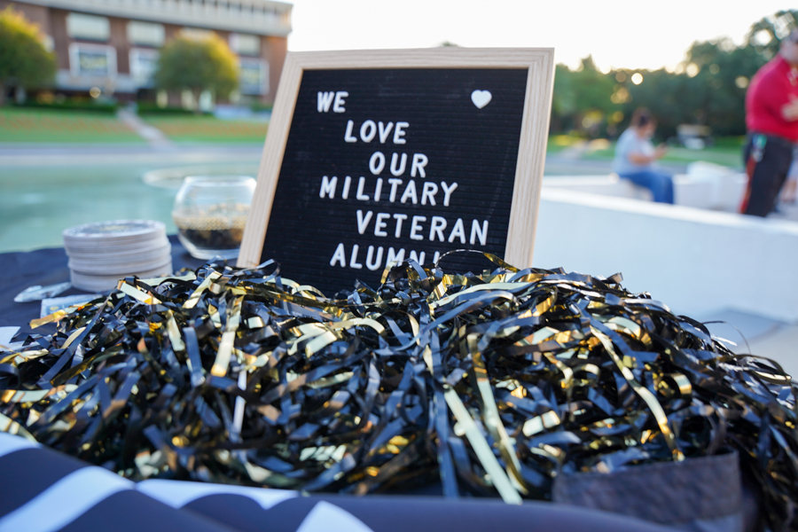 Framed sign reading 'We love our military veteran alumni' beside a pile of black and gold ribbons at an outdoor event on a campus-style setting