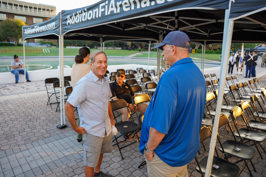 Two men talking under a blue canopy at an outdoor event; folding chairs are arranged in rows behind them.