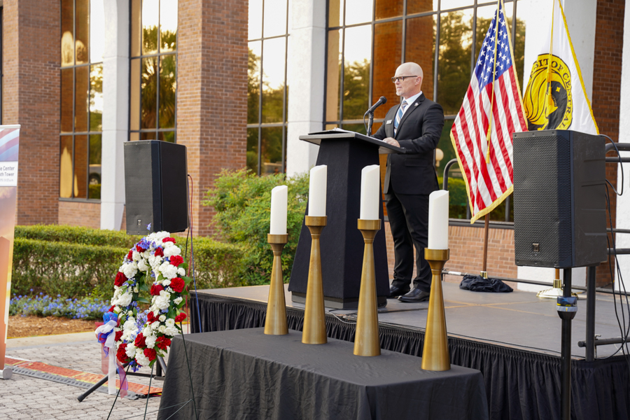 Man in a suit speaks at a podium on an outdoor ceremony stage, with the American flag and another flag nearby, and a floral wreath with candles on display beside him.
