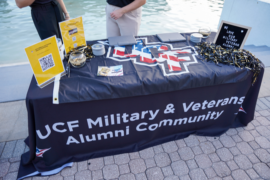 Promotional table for UCF Military & Veterans Alumni Community with flyers, QR codes, and black-gold decorations by the water.