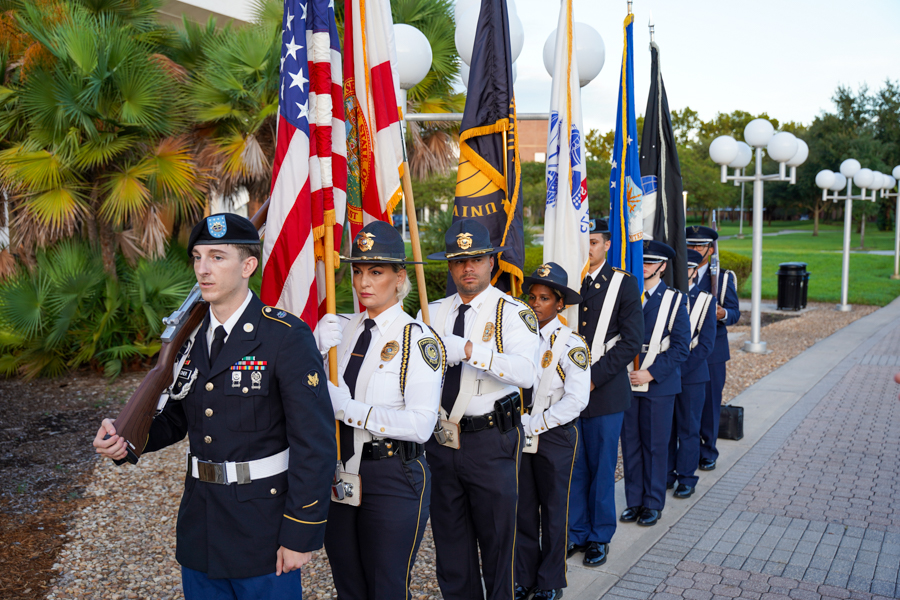 Color guard marching outdoors with multiple flags, led by uniformed personnel in formal dress.