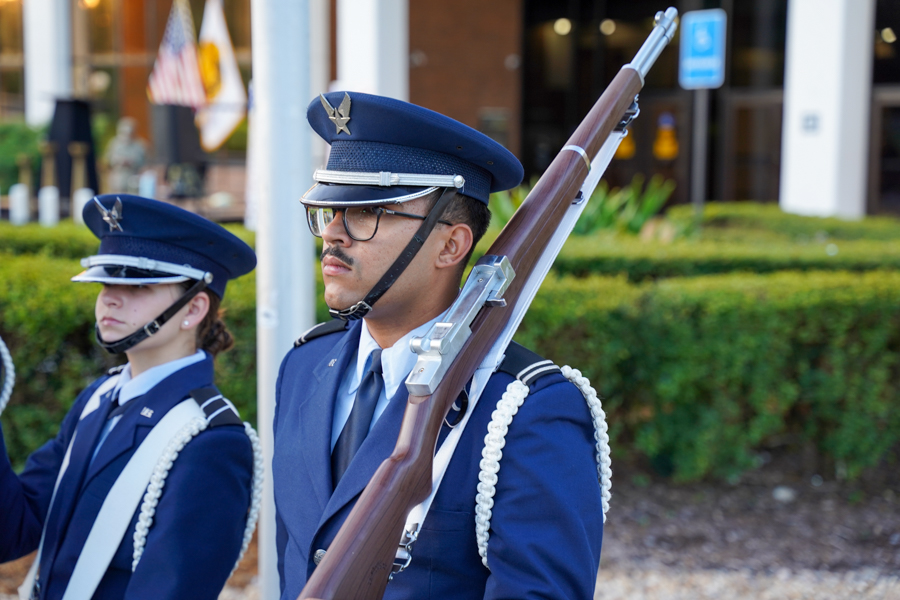 Ceremonial cadet march: two uniformed cadets in blue dress uniforms with rifles slung over shoulders, outdoors near a building.